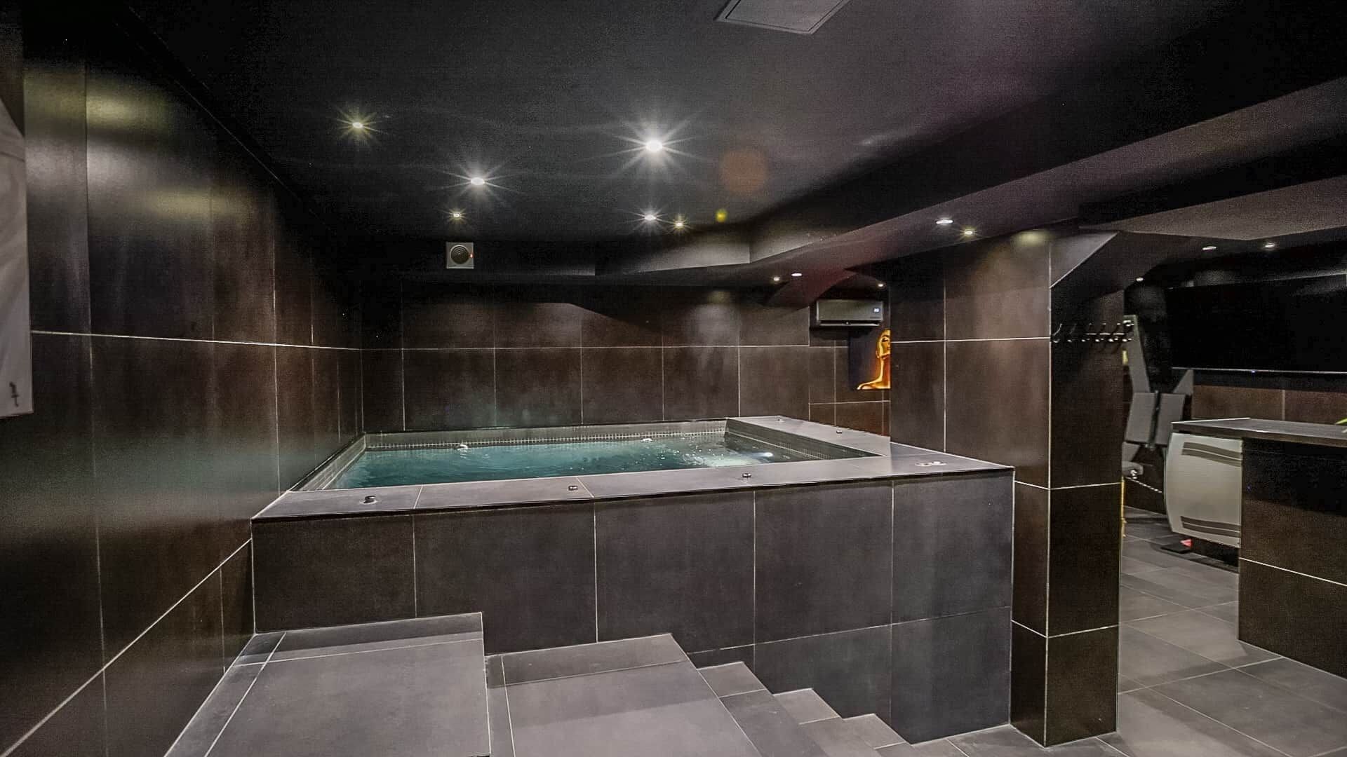 Une salle de spa moderne dotée d’un jacuzzi, de murs recouverts de carreaux foncés et d’un escalier menant à un niveau supérieur.