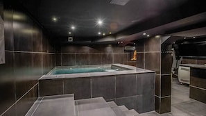 A modern spa room with a hot tub, dark tiled walls, and a staircase leading to a higher level. - Love Room—Jacuzzi Area / Massage Table (L'Escarène)