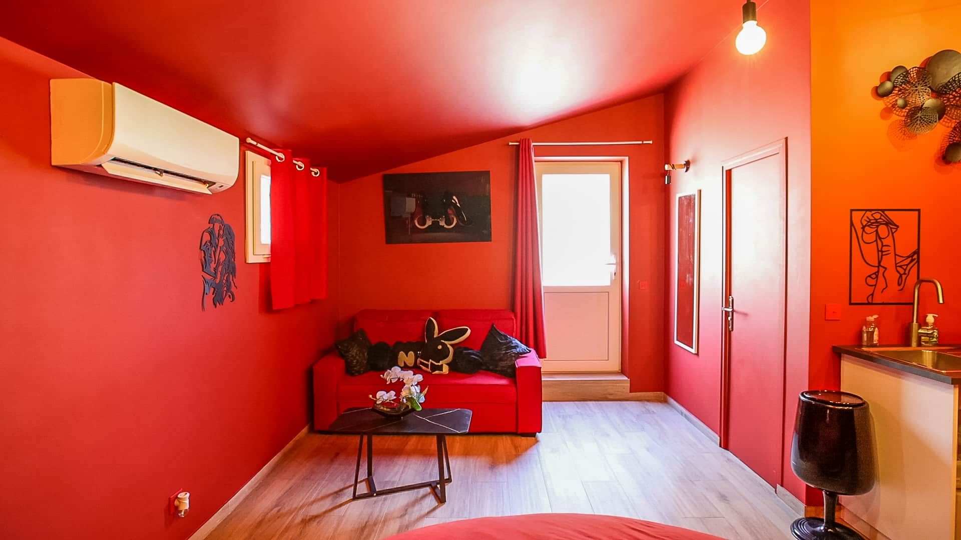 Een knus woongedeelte met een rode muur, een rode bank, een klein tafeltje met een vaas, een deur naar een andere kamer en een luchtverkoeler aan de muur.