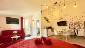A modern living room with a red sofa, a small round table with a vase, and a large red rug. There is a dining area with white chairs and a small table. - Love Room—Air-Conditioned Jacuzzi Area / Massage Table (L'Escarène)