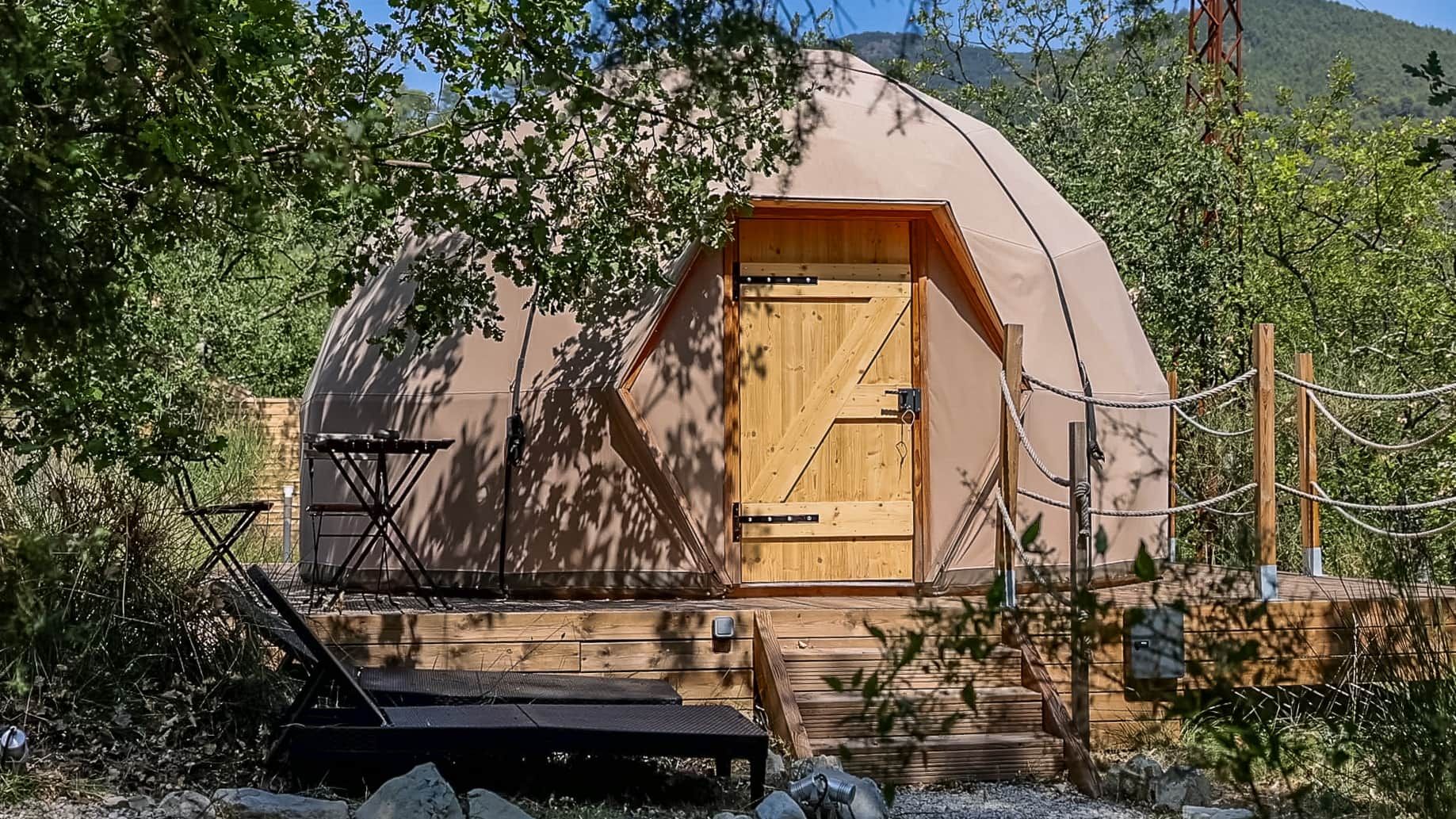 Une tente en forme de dôme avec une porte en bois, entourée d’arbres et dotée d’une terrasse en bois.