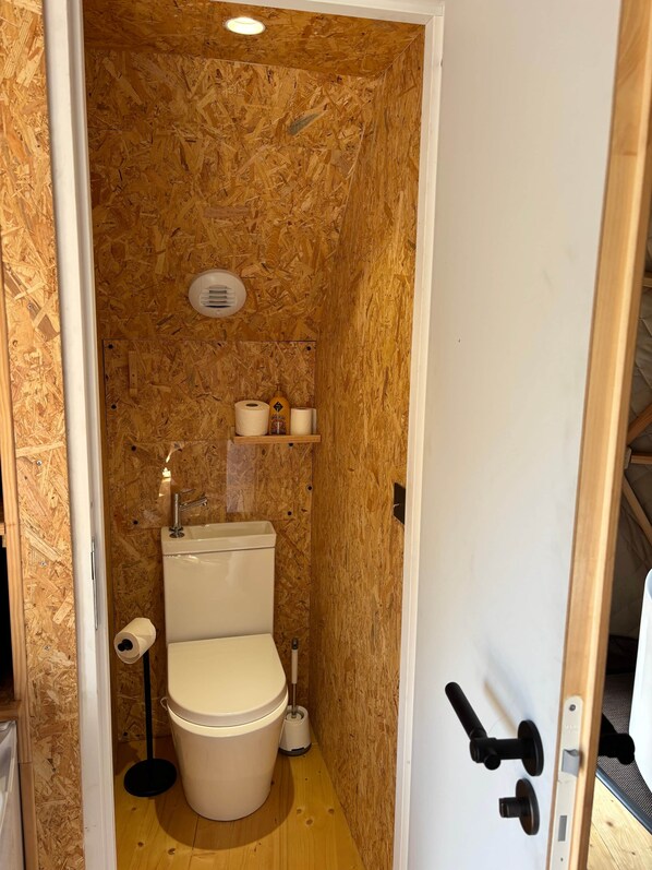 A small, compact bathroom with a wooden wall, a toilet, a sink, and a shelf with toiletries.