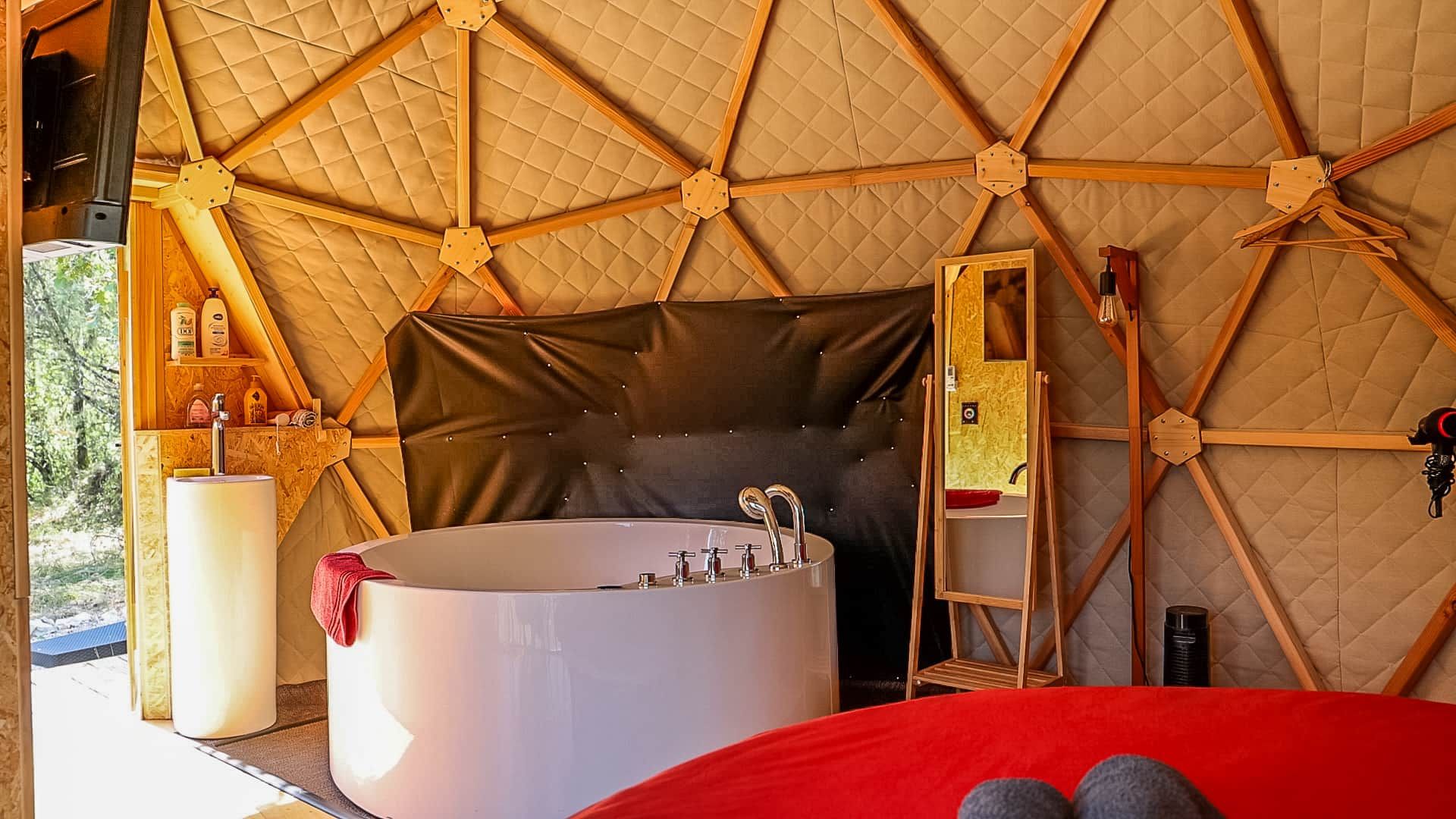 A tented accommodation with a large bathtub, a mirror, and a red bed.