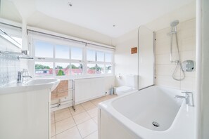 Une salle de bain moderne équipée d’une baignoire, de toilettes et d’un lavabo. Une fenêtre laisse entrer la lumière naturelle et offre une vue sur les bâtiments environnants.