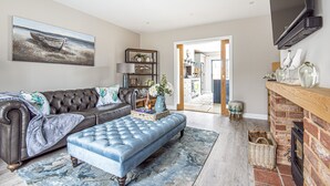 A living room with a leather sofa, a blue ottoman, a fireplace, and a television mounted on the wall. - Olive's Cottage - sleeps 6 guests  in 3 bedrooms (Burnham Market)