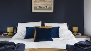 Un letto rifatto con lenzuola bianche, cuscini blu e dorati, e una coperta marrone. Due abat-jour con paralumi gialli, una sveglia digitale e un quadro incorniciato con una spiaggia.