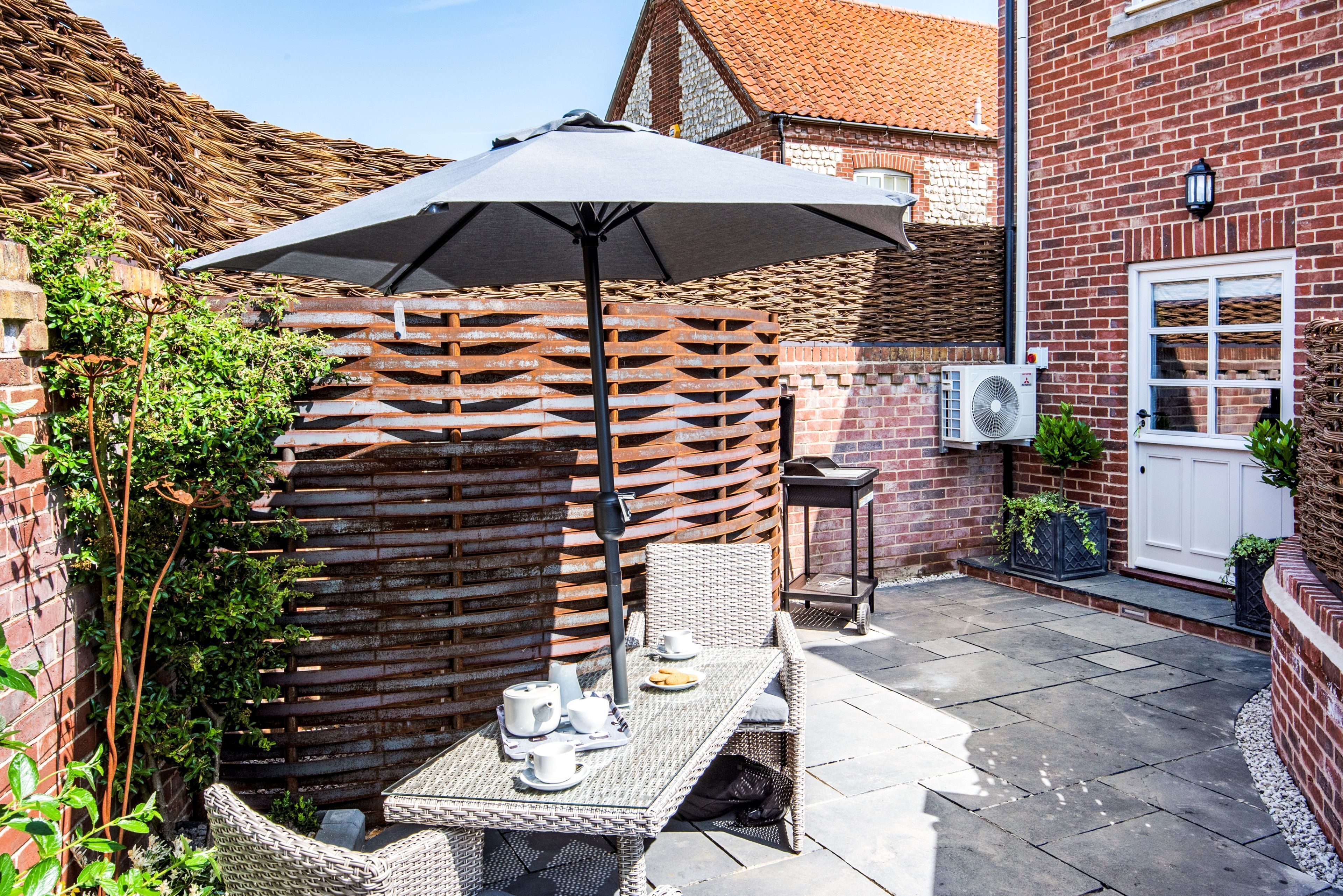 Une terrasse avec une table préparée pour le thé, une chaise en osier, une clôture en métal et un mur en briques.