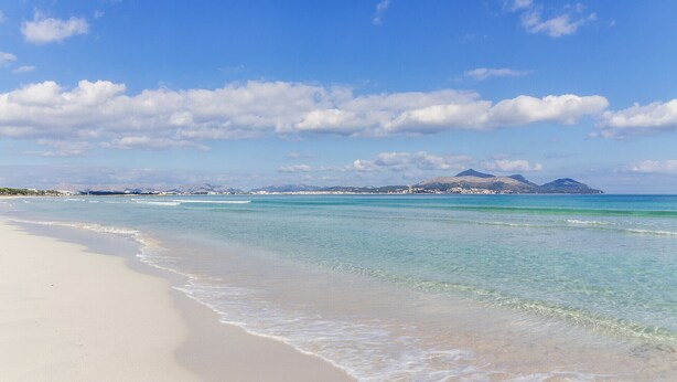 A beach with clear turquoise waters, a sandy shore, and distant mountains under a partly cloudy sky. - Can Segue by Balearic Villas (Alcúdia)