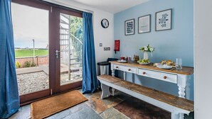 A room with a wooden bench, a white dresser, a dining table with a vase of flowers, and a view through a glass door to an outdoor area with a fence and greenery. - Longview Cottage - sleeps 6 guests  in 3 bedrooms (Wells-next-the-Sea)