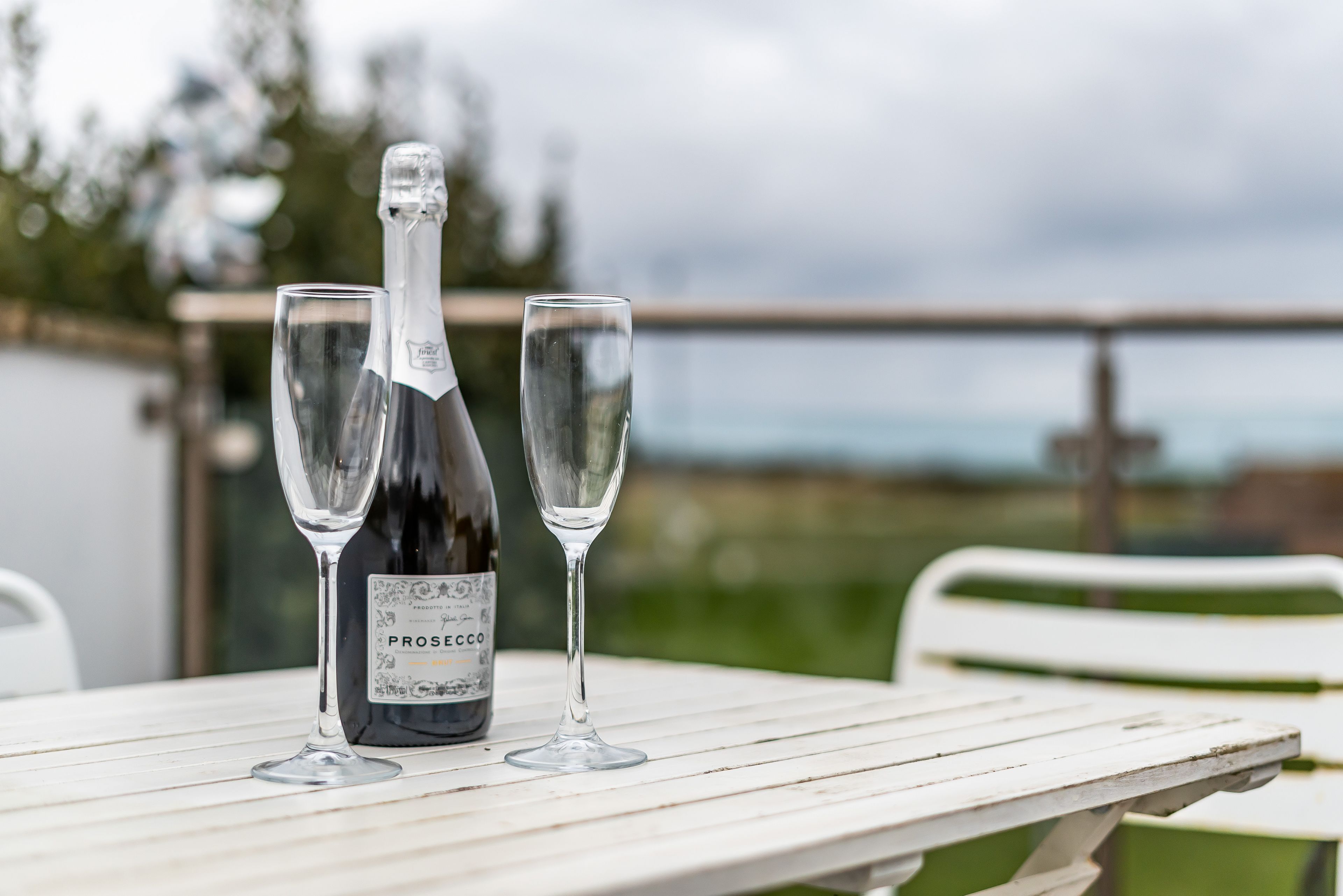 A bottle of Prosecco wine and two glasses on a white table with a view of the outdoors.