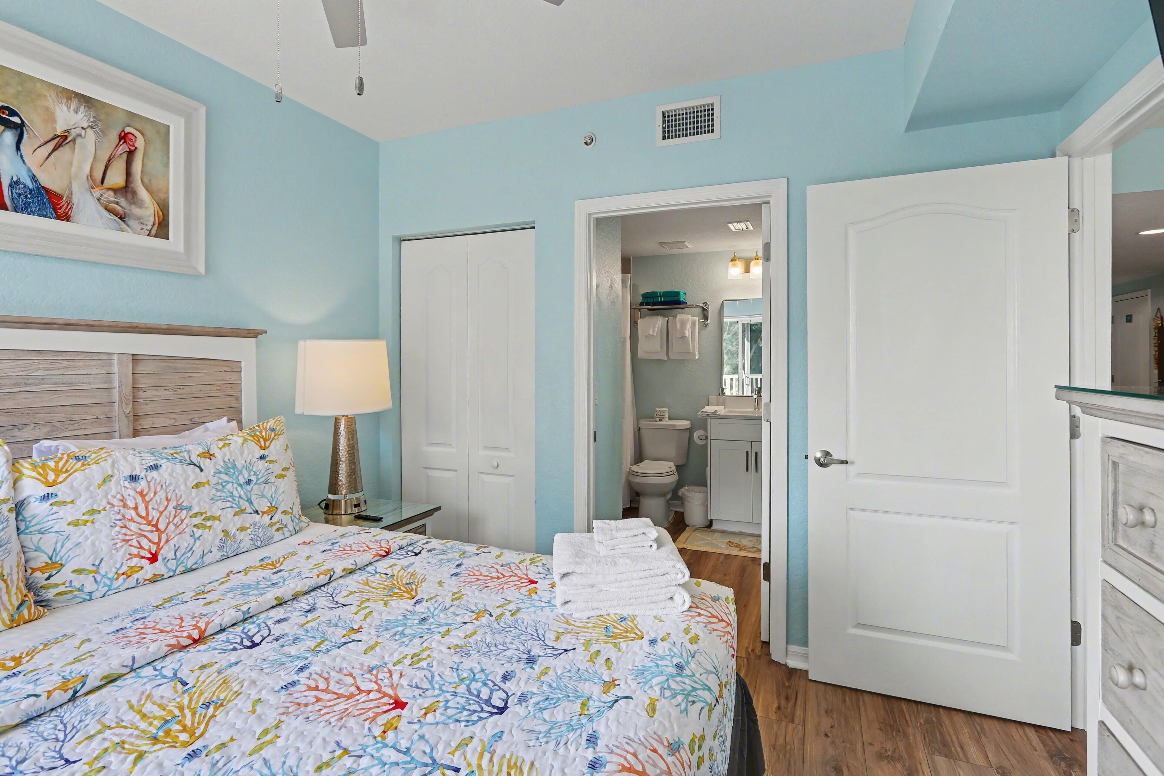 A bedroom with a bed, a nightstand, a lamp, a painting, and a bathroom visible through an open door.
