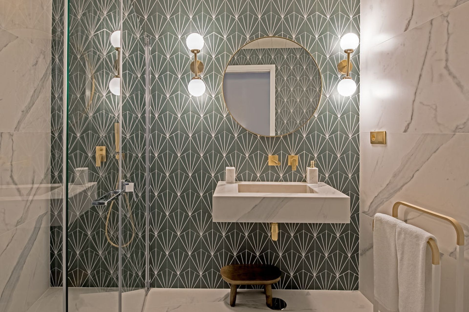 Une salle de bain avec un mur orné d’un motif géométrique, un lavabo blanc, un miroir rond et deux appliques murales.