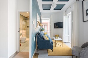Uma sala de estar moderna com um sofá azul, uma mesinha e um tapete amarelo. É visível um banheiro ao fundo, com chuveiro e vaso sanitário.