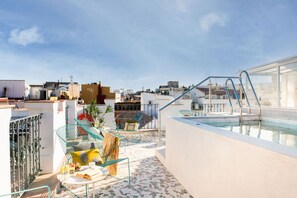 Uma área de piscina no terraço, com guarda-corpo de vidro, espaço de estar com mesa e cadeiras, e vista para a cidade.