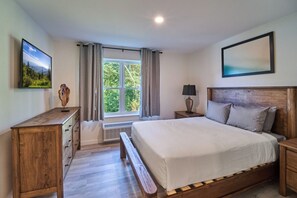 Ein Schlafzimmer mit einem hölzernen Bett, einem Nachttisch, einem Tisch am Bett, einem Fernseher, einem Fenster mit Vorhängen und einem Gemälde an der Wand.