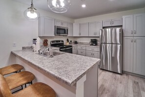 A modern kitchen with white cabinets, a stainless steel refrigerator, a microwave, and a granite countertop. There are three brown bar stools and a hanging light fixture. - Ruby 215: 2BD | Great Location + Shared Amenities! (Pigeon Forge)