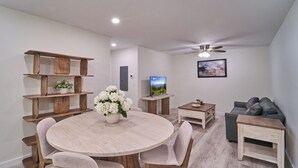 Una sala moderna con una mesa de comedor redonda de madera, sillas de color claro, un sofá, un televisor y un ventilador de techo.
