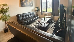 Un salon moderne avec un canapé en cuir noir, une table basse et un téléviseur. Il y a une fenêtre avec des rideaux, une plante et un tableau au mur.