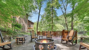 A fire pit surrounded by wooden chairs and tables in a forested area with a stone building in the background. - River's Edge Retreat (Sevierville)
