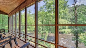 A covered outdoor seating area with wooden benches and large windows offering a view of a river and forest. - River's Edge Retreat (Sevierville)