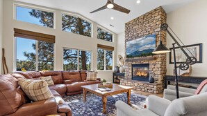 A spacious living room with a stone fireplace, leather sofa, and large windows offering a view of trees. - NEW Cabin | Pool Table | Golf Course | AC | EV Charger (Flagstaff)