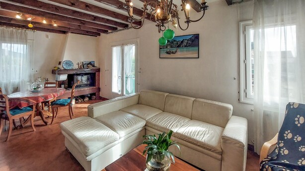 Ein Wohnzimmer mit beiger Couch, einem Esstisch mit Stühlen, einem Kamin und einer Chandelüster.