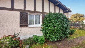 Ein Haus mit weißer Fassade, dunklen Holzbalken, einem Fenster mit weißen Vorhängen und einem gepflegten, grünen Heckenstreifen.