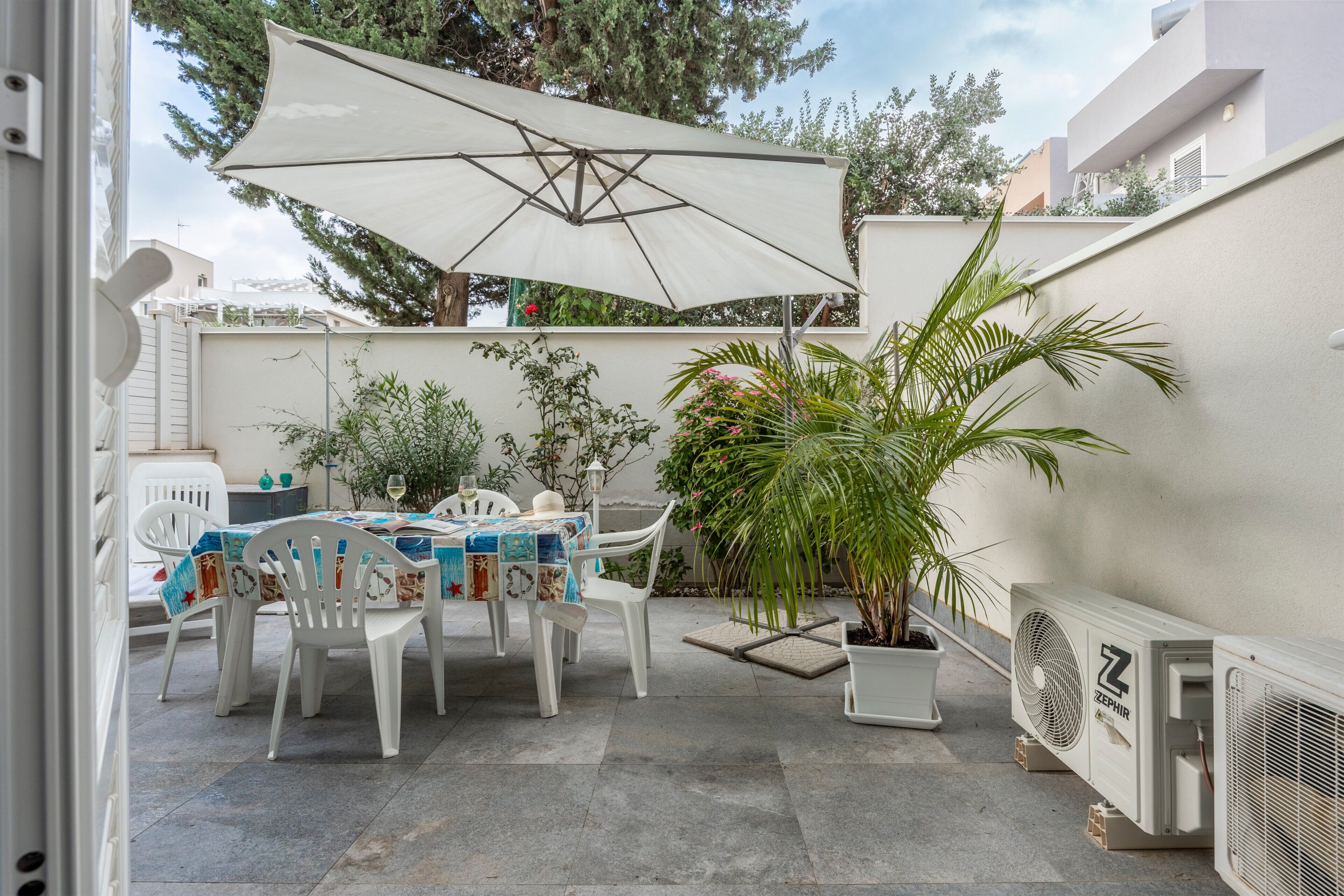 An outdoor patio with a table, chairs, a large umbrella, potted plants, and an air conditioning unit.