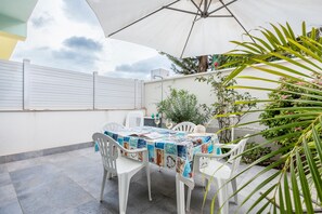 Een terras met een tafel, bedekt met een kleurrijke tafelkleed, omringd door witte plastic stoelen, onder een grote parasol.