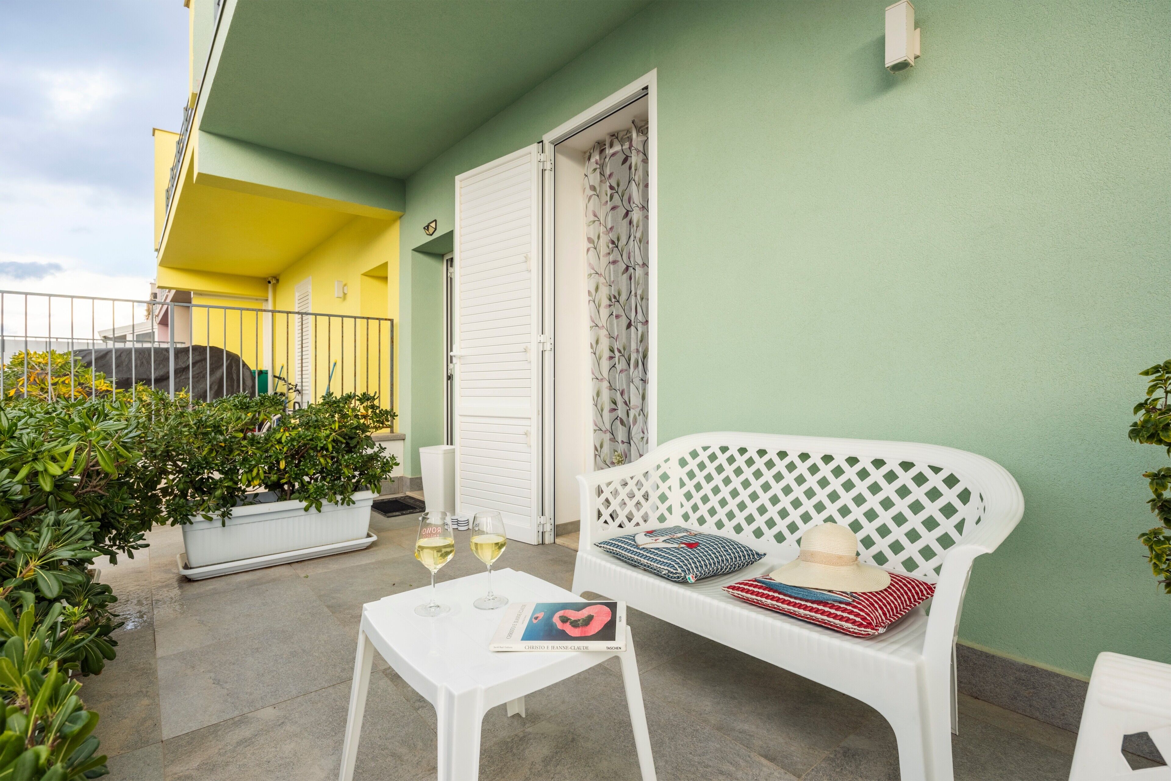 Un balcon avec une table blanche, deux verres de vin, un livre, un banc blanc avec un coussin à motifs et une plante en pot.