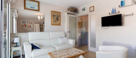 Una sala de estar con un sofá blanco, una mesa de centro de madera y un sillón blanco. Hay un televisor montado en la pared y un cuadro a la vista.