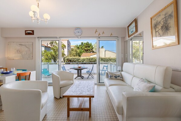 A bright living room with a white sofa, a wooden coffee table, and a dining table with chairs. There is a balcony with a view of the neighborhood.