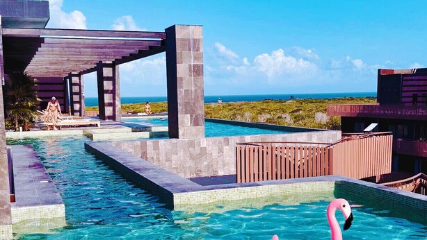 Una piscina azul cristalina con un flotador de flamenco rosa, rodeada por una terraza de madera y una construcción de piedra.