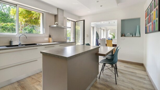 Una cucina moderna con isola centrale, zona pranzo con tavolo e sedie, e vista sul verde all'esterno.