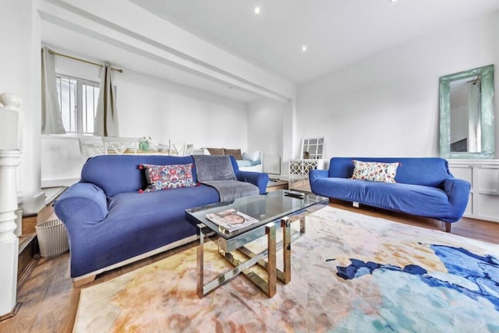 A spacious living room with two blue sofas, a glass coffee table, and a large abstract rug.
