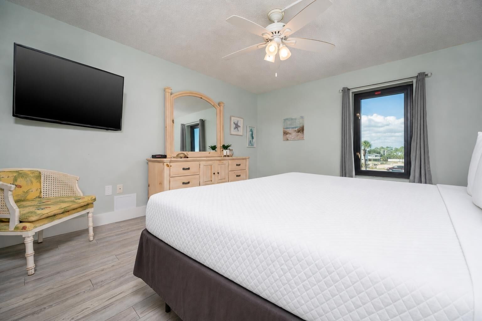 A bedroom with a bed, a chair, a dresser, a mirror, a ceiling fan, and a window with a view.