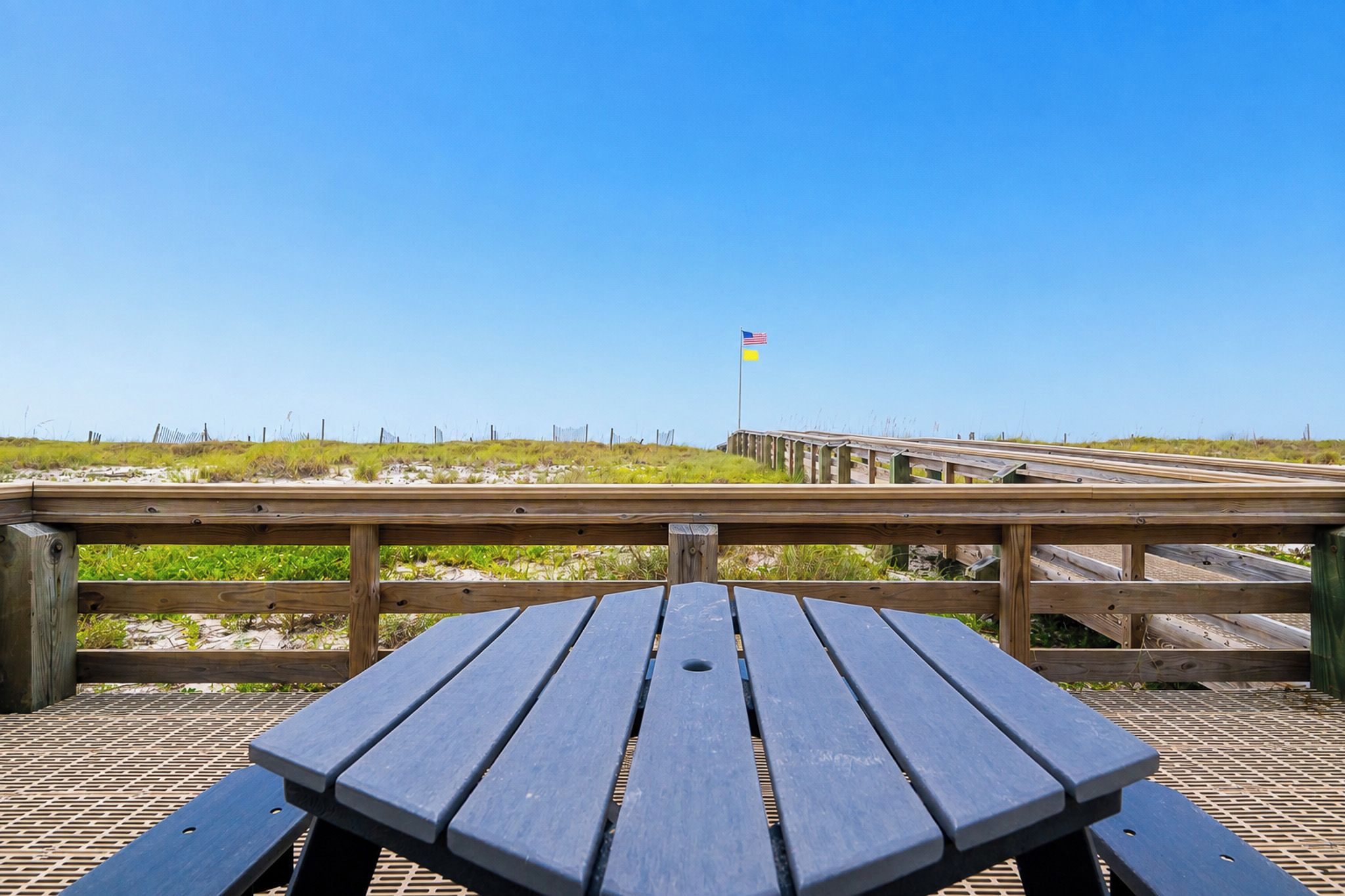 Une table de pique-nique en bois avec un plateau en métal ajouré, une terrasse en bois dotée d’une rambarde et un mât sur lequel flotte un drapeau, le tout sur fond de paysage.