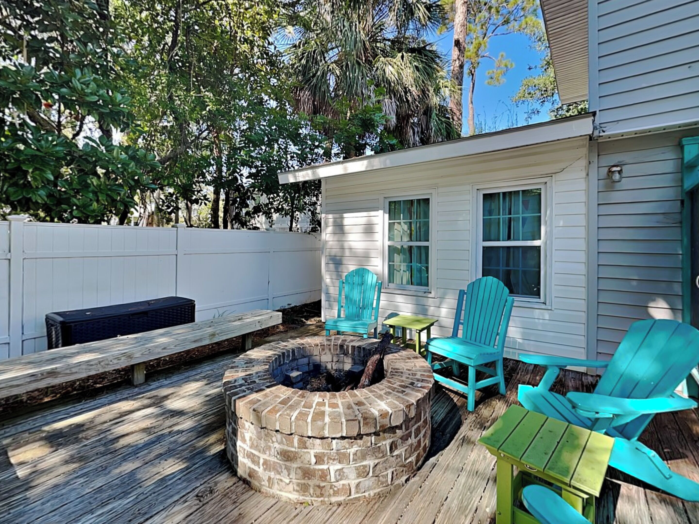 A backyard with a brick fire pit, wooden deck, and turquoise Adirondack chairs.