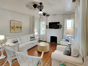 A spacious living room with a white fireplace, a television mounted above it, and a ceiling fan. There are two white sofas and a wicker chair. - Coast A While (Tybee Island)