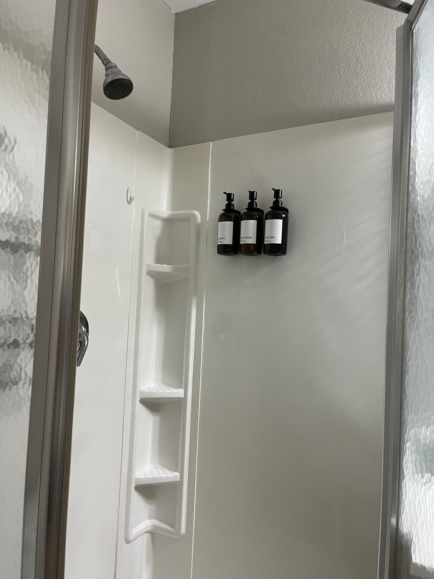 Zona de ducha con mampara de vidrio, cabezal de ducha y tres botellas de champú y acondicionador en la pared.