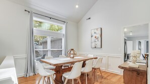 Un comedor con mesa de madera y sillas blancas, una ventana con cortinas, y un cuadro en la pared.