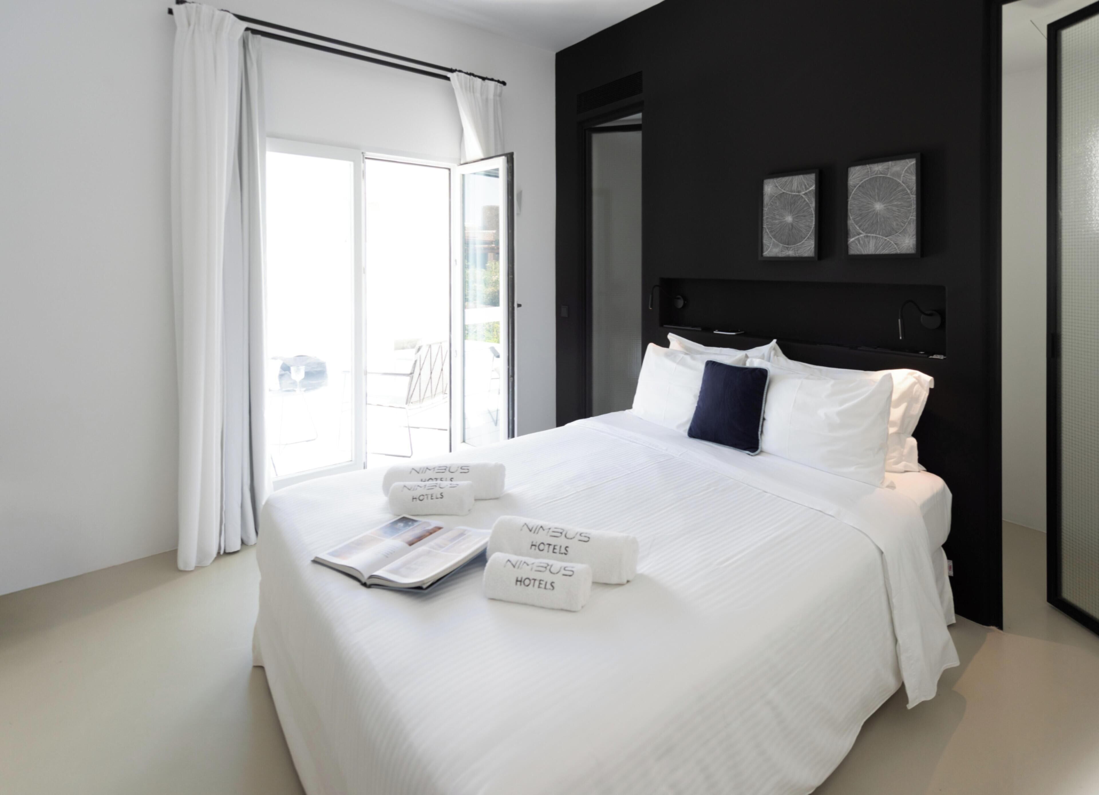 A modern hotel room with a large bed, white bedding, a magazine on the bed, and a view of the outdoors through the window.