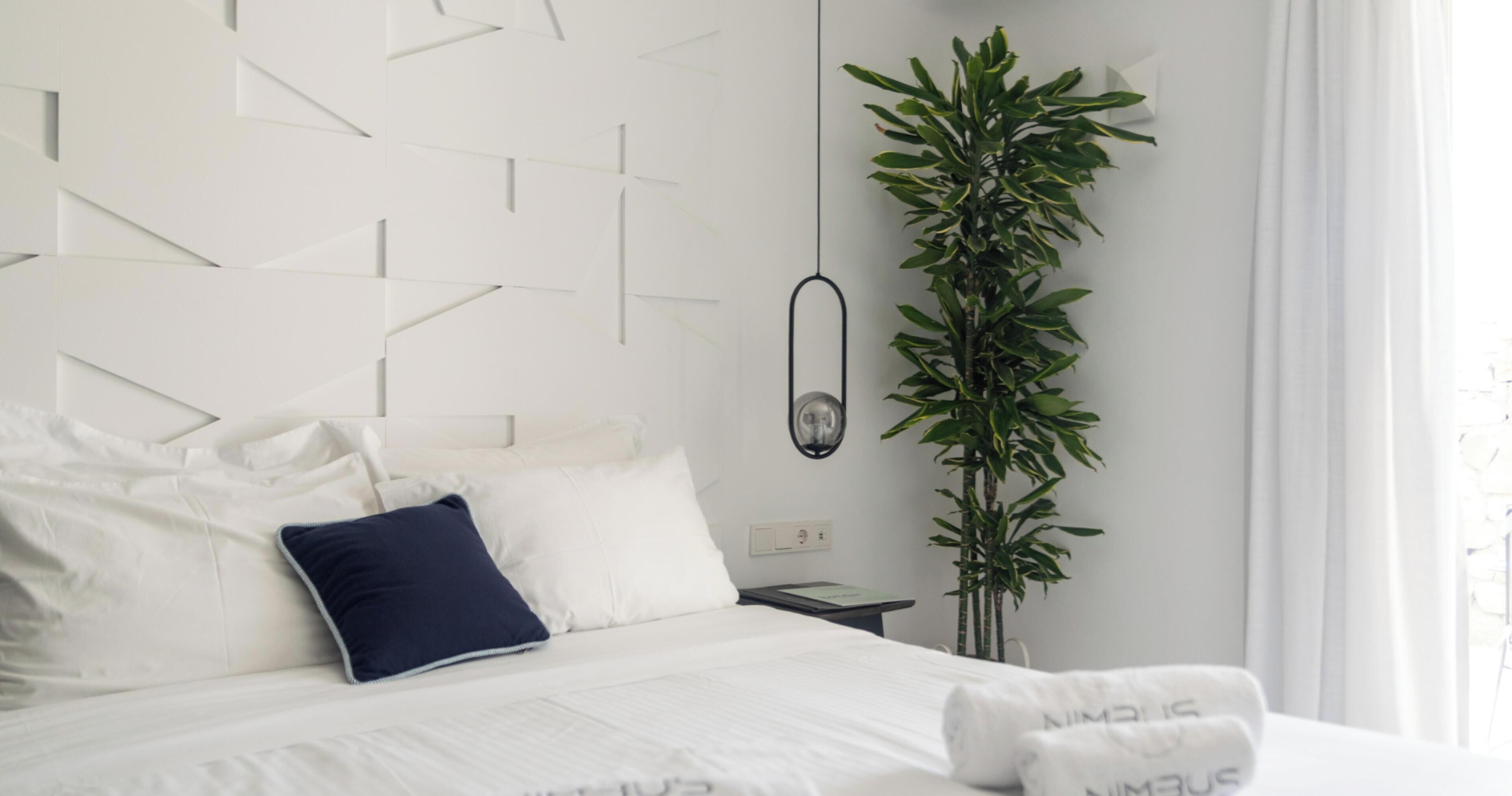 A modern hotel room with a geometric-patterned headboard, a large potted plant, and neatly made bed with white linens and a dark blue pillow.