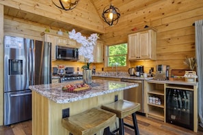 A modern kitchen with wooden cabinetry, stainless steel appliances, a granite countertop, and a central island with bar stools. - Ridge Hollow Retreat – Hot Tub, Fire Pit, Dogs (Gatlinburg)