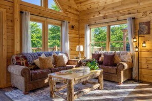 A cozy cabin interior with two sofas, a coffee table, and large windows offering a view of the outdoors. - Ridge Hollow Retreat – Hot Tub, Fire Pit, Dogs (Gatlinburg)