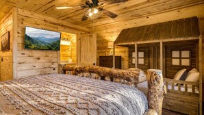 A log cabin bedroom with a bed, a ceiling fan, a mounted TV, and a rustic wooden bench. - Ridge Hollow Retreat – Hot Tub, Fire Pit, Dogs (Gatlinburg)