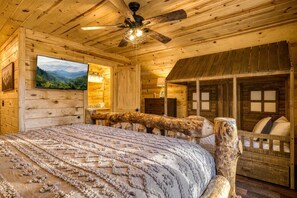 Ein Schlafzimmer in einer Blockhütte mit einem Bett, einem Deckenventilator, einem festmontierten Fernseher und einer rustikalen Holzbank.