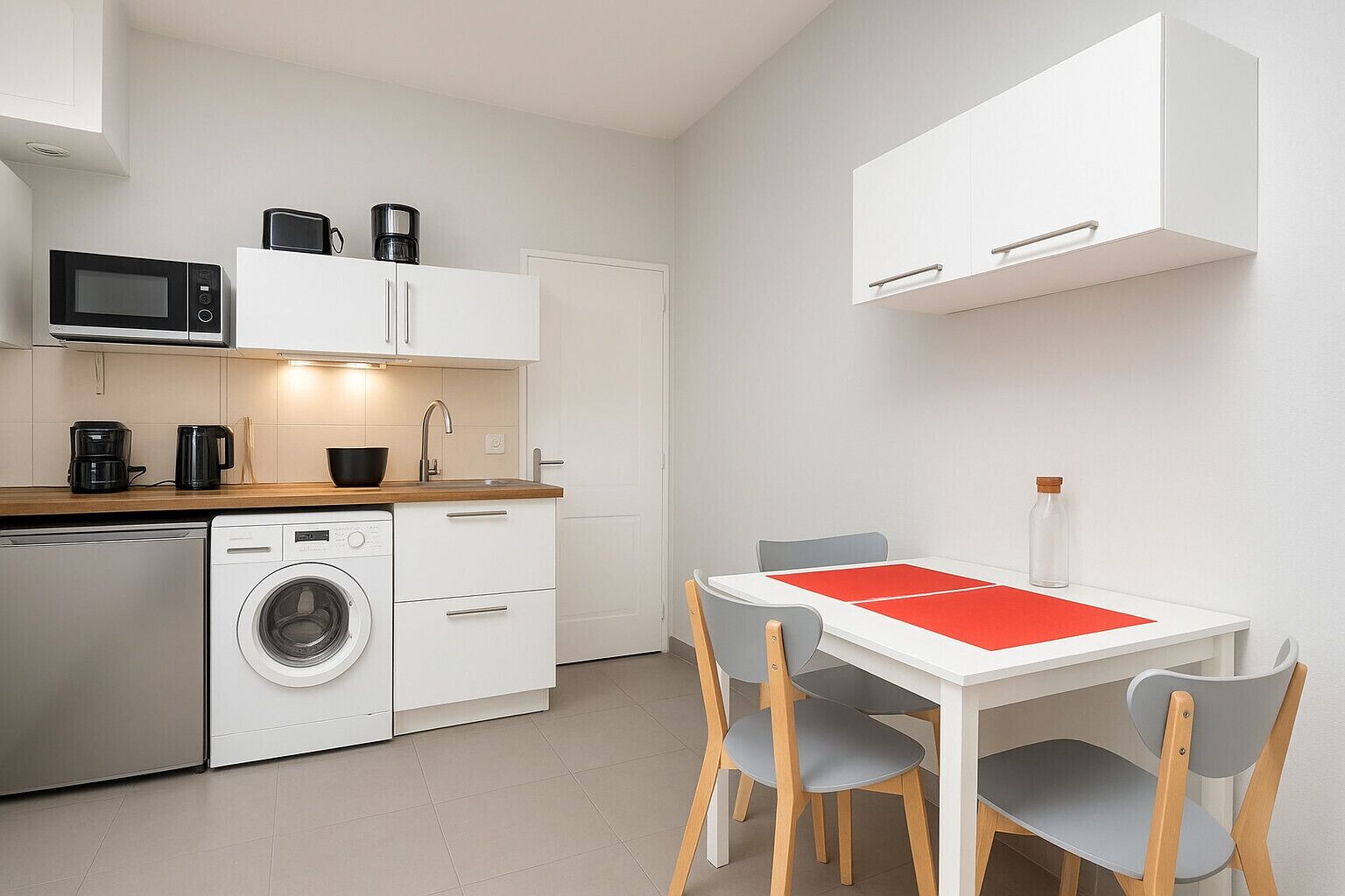 Cocina compacta con lavadora, microondas y cafetera. Hay una mesa de comedor con sillas y un mantel individual rojo.