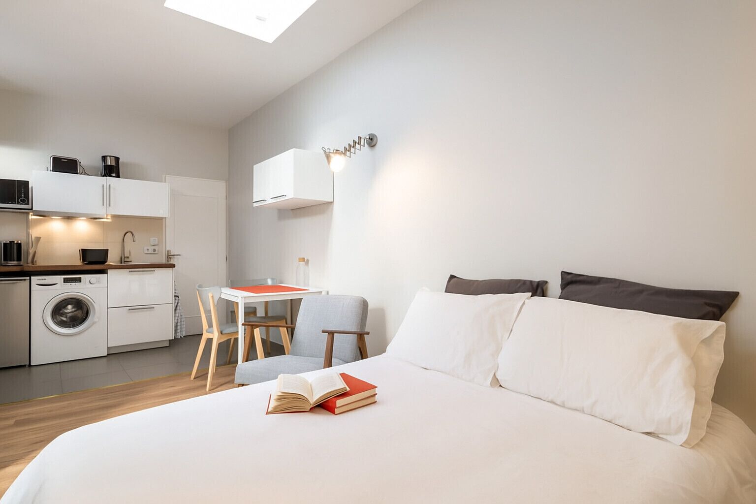 Een moderne slaapkamer met een groot bed, een klein bureau en een kitchenette op de achtergrond.