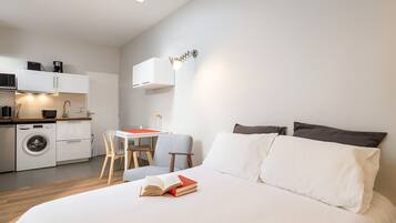 Een moderne slaapkamer met een groot bed, een klein bureau en een kitchenette op de achtergrond.